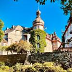 Schloss mit Turm und Kletterpflanzen, umgeben von Bäumen und Steinmauern unter blauem Himmel.