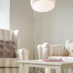 Ein Wohnzimmer mit Sessel und Sofa, Beistelltisch und Stehlampe. Fenster mit Blick nach draußen.