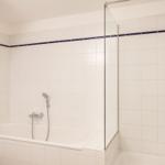 Bad mit Badewanne und Dusche, weiße Fliesen, dunkelblaue Zierleiste, Holzbalken an der Decke.
