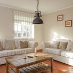 Zwei Sofas mit Streifenbezug, Holztisch und Wandbilder in einem Wohnzimmer mit Fenstern und Terrakottaboden.