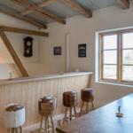 Raum mit Holzbar, Fellhockern und Fensterblick. Deckenbalken und Uhr sichtbar.