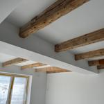 Raum mit sichtbaren Holzbalken an der Decke und grauen Wänden. Fenster mit gelber Schutzfolie.