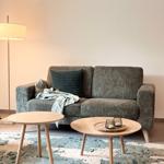 Ein grauer Sofa mit Kissen und Decke, zwei runde Holztische und ein Stehlampe.