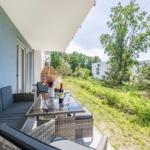 Terrasse mit Sitzgruppe, Tisch und Weinflasche. Grünfläche und Bäume im Hintergrund.
