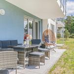 Terrasse mit Sitzgruppe, Esstisch und Hängesessel. Grünfläche und Bäume im Hintergrund.