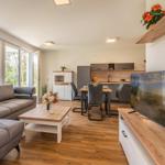 Wohn- und Essbereich mit Sofa, TV und Küchenzeile. Holzboden und große Fenster.