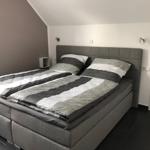 Ein Schlafzimmer mit einem großen Bett und grauer Bettwäsche. Die Wände sind hell und die Decke ist schräg.