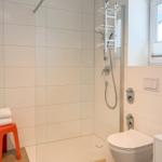 Dusche mit Duschbrause, Toilette und Waschbecken. Ein roter Hocker steht neben dem Heizkörper.