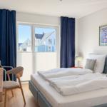 Schlafzimmer mit Bett, Schreibtisch, TV und Fensterblick auf Häuser.