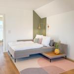 Schlafzimmer mit Bett, Teppich und Wandbeleuchtung. Tür führt zu einem Balkon.