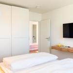 Schlafzimmer mit Bett, Kleiderschrank und Wand-TV. Tür führt in einen Flur.