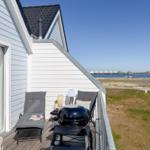 Terrasse mit Liegestühlen und Grill, Blick auf Wasser und Strand.