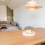 Esszimmer mit Holztisch und Sofa im Hintergrund. Deckenleuchte und Wandbild sichtbar.