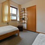 Zimmer mit zwei Betten, Fenster, Bücherregal und Tür.