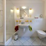 Badezimmer mit Dusche, Waschbecken und Toilette. Gelbe Halterungen und ein gelb-weißes Badetuch.
