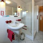 Badezimmer mit Dusche, Waschbecken und Spiegel. Rote Haltegriffe und Handtuchhalter.