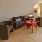 Wohnzimmer mit Sofa, zwei Sesseln, Tisch und Fernseher. Holzboden und Fenster mit Vorhängen.