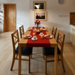 Esszimmer mit Holztisch, sechs Stühlen und roter Tischdecke. Wandbilder und Uhr sind sichtbar.