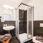 Badezimmer mit Dusche, Waschbecken und Toilette. Fliesen und Holzstuhl sichtbar.