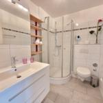 Badezimmer mit Dusche, Waschbecken und Toilette. Weiße Fliesen und Holzregale.