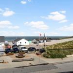 Parkplatz mit Gebäuden und Strand im Hintergrund unter blauem Himmel.