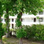 Weißes Mehrfamilienhaus mit Balkonen, umgeben von Bäumen und Sträuchern.