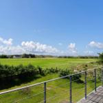 Terrasse mit Blick auf grünes Feld und Häuser unter blauem Himmel.