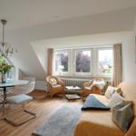 Wohn- und Essbereich mit Sofa, Stühlen und Fenstern. Holzboden und Deckenlicht.