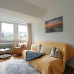 Wohnzimmer mit Sofa, zwei Sesseln und großen Fenstern. Wandbild zeigt Strand bei Sonnenuntergang.
