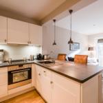 Küche mit Arbeitsplatte, Herd und Spüle. Im Hintergrund Wohnbereich mit Sofa und Fenster.
