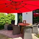 Terrasse mit runden Tisch, vier Stühlen und rotem Sonnenschirm. Hintergrund: grüne Hecke und Fenster.