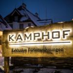 Kamp Hof: Exklusive Ferienwohnungen. Holzschild mit Beleuchtung vor schneebedeckten Gebäuden.