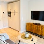 Wohnzimmer mit Holzboden, Fernseher und Sideboard. Küchenbereich mit Hochschrank sichtbar.