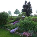 Garten mit Blumenbeeten, Sträuchern und einem Pavillon. Hintergrund mit Bäumen.