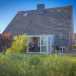 Haus mit Glasveranda, Garten und Satellitenschüssel. Personen sitzen im Innenbereich.