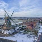 Schneebedeckte Landschaft mit historischem Windrad und Häusern entlang einer Straße.