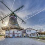 Windmühle mit angrenzendem Gebäude und Gartenbereich unter blauem Himmel.