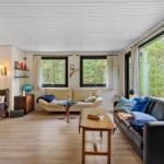 Wohnzimmer mit Sofa, Couch und Tischen. Große Fenster mit Blick auf Wald.