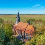 Kirche mit Turm und rotem Dach umgeben von Bäumen und Feldern unter blauem Himmel.