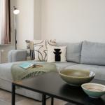Ein grauer Sofa mit Kissen und einer Decke. Auf dem Tisch stehen Schalen und ein Glas.