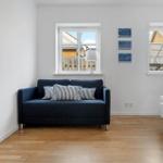 Ein Raum mit Sofa, Bett und zwei Fenstern. Holzfußboden und weiße Wände.