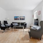 Wohnzimmer mit schwarzer Couch, zwei Sesseln und Holzboden. An der Wand hängen zwei Bilder.