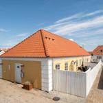 Haus mit rotem Dach und gelber Fassade, umgeben von weißem Zaun und gepflastertem Hof.