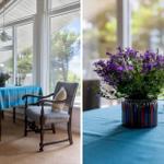 Esstisch mit blauer Tischdecke und Stuhl vor großem Fenster. Vase mit lila Blumen und Kerzenhaltern.
