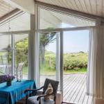 Ein Essbereich mit Tisch und Stühlen. Durch große Fenster sieht man einen Holzdeckenbereich und Grünland.
