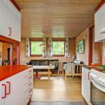 Küche mit rotem Arbeitsplatz und weißer Einbauküche. Wohnbereich mit Sofa und Fenstern.