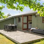 Holzterrasse mit Tisch, Stühlen und Liege vor einem Haus mit offener Tür.