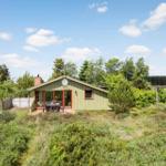 Ein kleines Haus mit Terrasse, umgeben von Grün und Bäumen unter blauem Himmel.