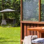 Terrasse mit Holztisch und Blick auf grünen Garten mit Sonnenschirm und Liegestühlen.