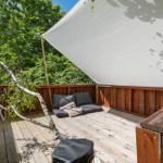 Holzterrasse mit Sitzbank, Sonnensegel und Baumast. Umgeben von Bäumen.